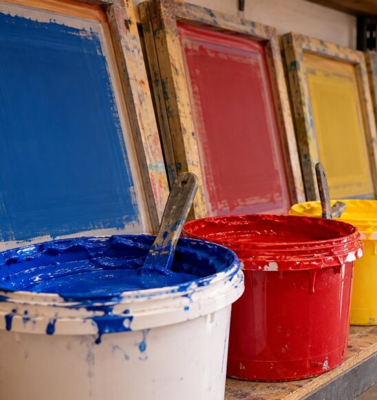 Buckets of blue, red, yellow and green screen printing ink in front of coated screens